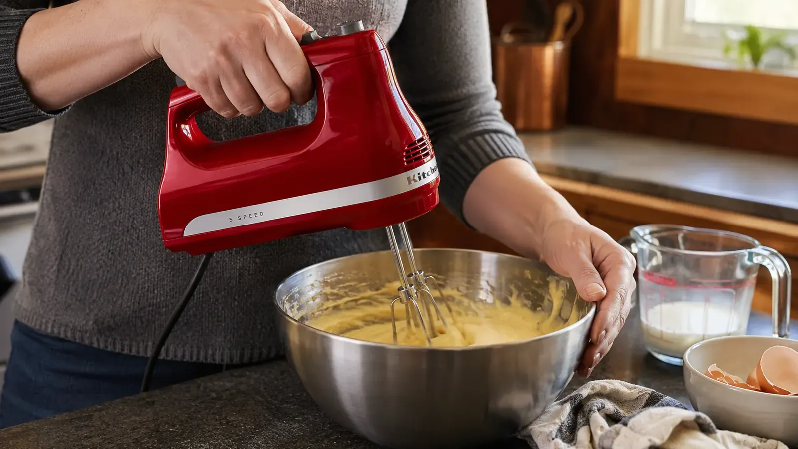 Hands holding a red KitchenAid hand mixer while beating batter in a stainless steel bowl