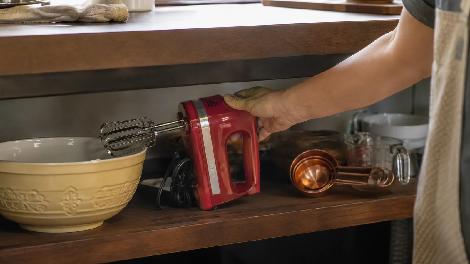 A hand mixer sitting on an open shelf next to measuring cups instead of tucked away in a drawer