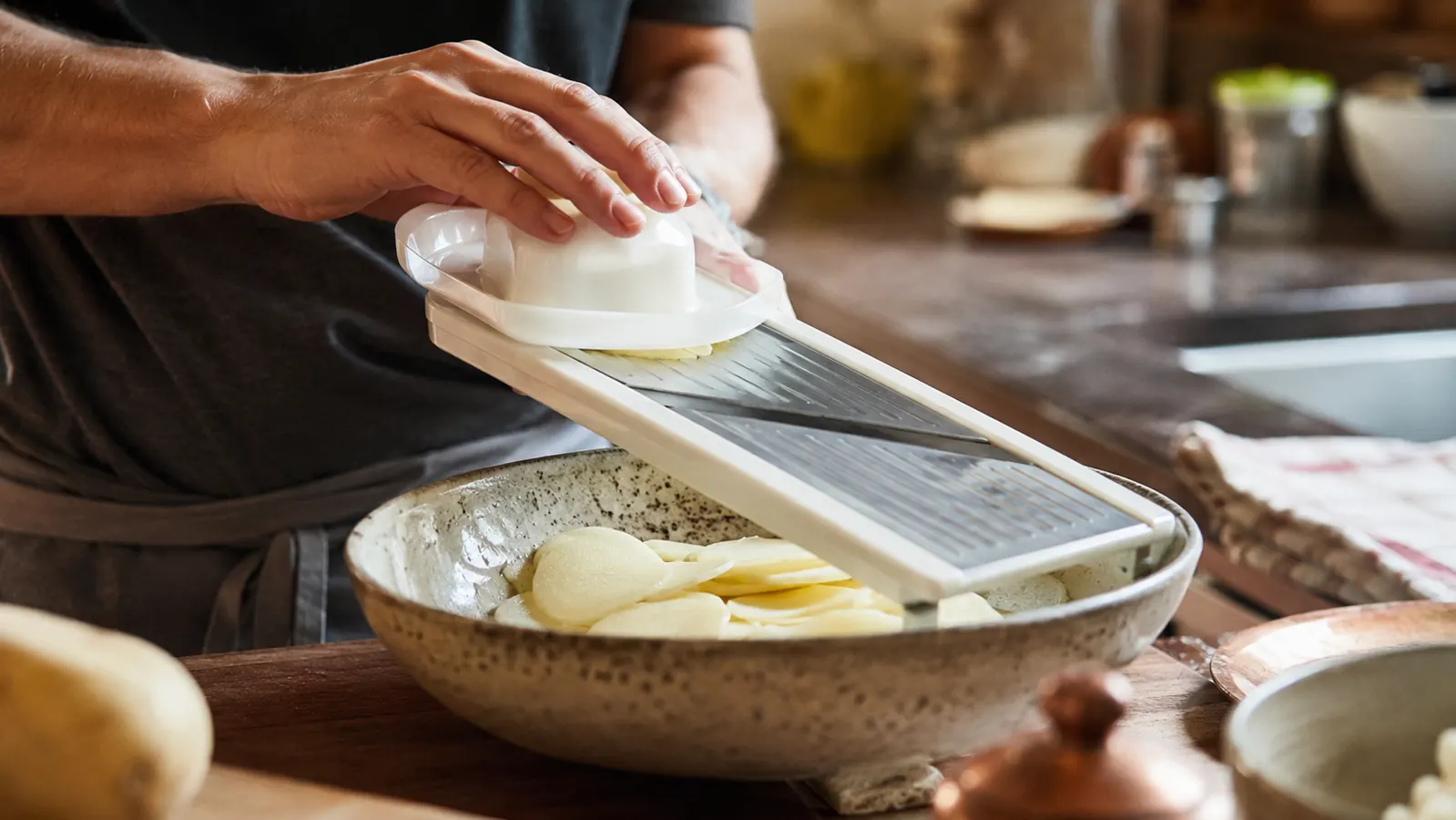 A hand using a mandoline slicer with a food holder to cut thin potato slices for a gratin